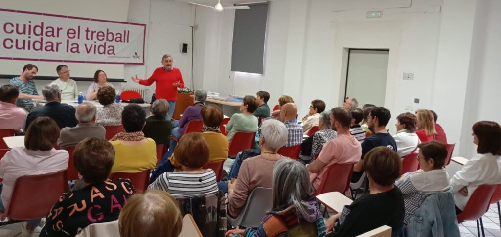 Speaker in a red sweater addresses a seated audience in a bright room, with a banner reading 'cuidar el treball cuidar la vida' on the wall.