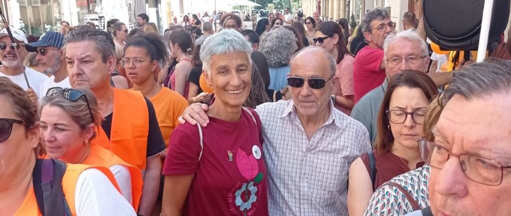 Crowd of people walking down a sunny street at an outdoor event; a smiling woman with gray hair in a maroon shirt stands near a man in sunglasses in the center.