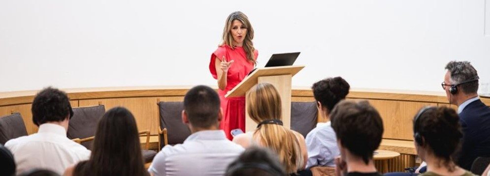 Speaker in a red dress stands at a wooden lectern addressing a seated audience in a conference room.