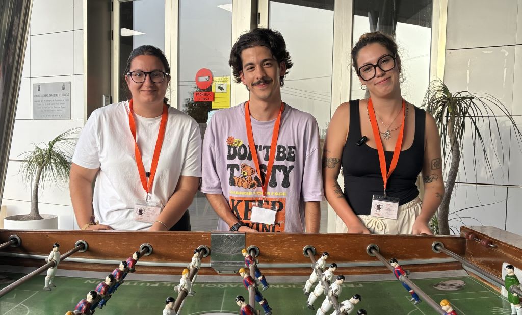 Three people with orange lanyards pose behind a foosball table, looking at the camera with smiles.