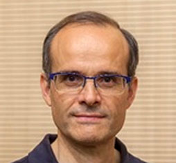 Head-and-shoulders portrait of a middle-aged man with short gray hair, blue glasses, and a dark polo, looking at the camera.