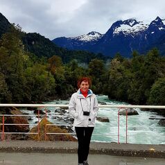 Person standing on a railing of a bridge beside a rushing river, with forest and snow-capped mountains in the background.