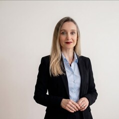 Professional woman with blonde hair in a black blazer and light blue shirt, standing against a plain white wall, hands loosely clasped.