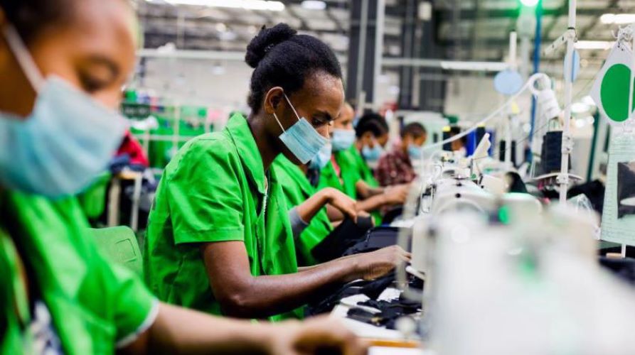 Factory workers in green uniforms and masks sewing on an assembly line in a garment factory.