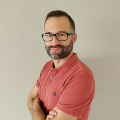 Portrait of a man with glasses and a beard, wearing a pink polo shirt with arms crossed against a light beige wall.