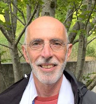 Smiling man with gray beard and glasses outdoors, wearing a red shirt and dark jacket, with trees in the background.