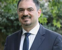 Portrait of a man in a navy suit and blue tie outdoors with greenery in the background.