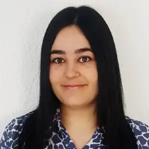 Portrait of a young woman with long dark hair, wearing a blue patterned blouse, smiling against a light wall.
