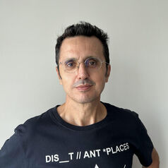 Portrait of a man with short dark hair and glasses, wearing a black T-shirt against a light wall, facing the camera.