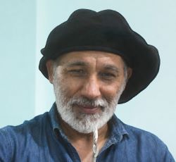 Portrait of an older man with a gray beard wearing a black beret-style hat and blue denim shirt, facing the camera.