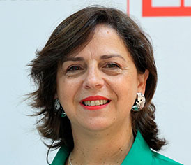 Portrait of a smiling woman with short dark hair wearing a green blazer and turquoise earrings against a light background.