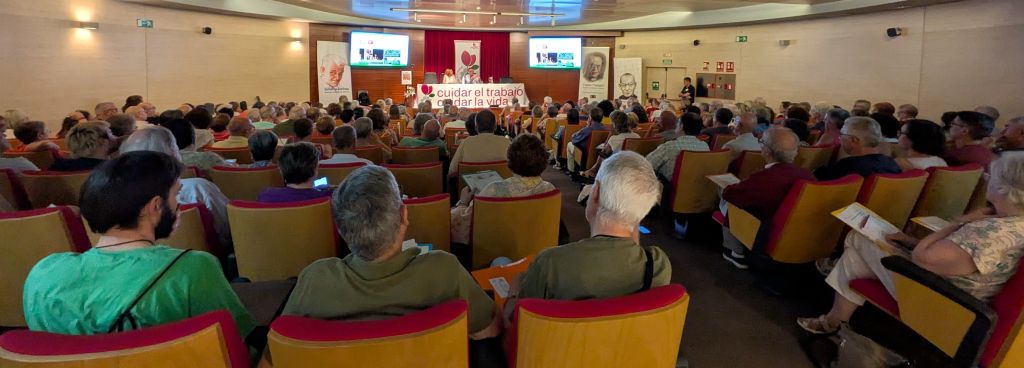 Large auditorium filled with people watching a panel at the front; banners and screens behind the speakers on stage.