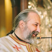 Bearded priest in white and gold vestments speaking into a microphone in profile view.