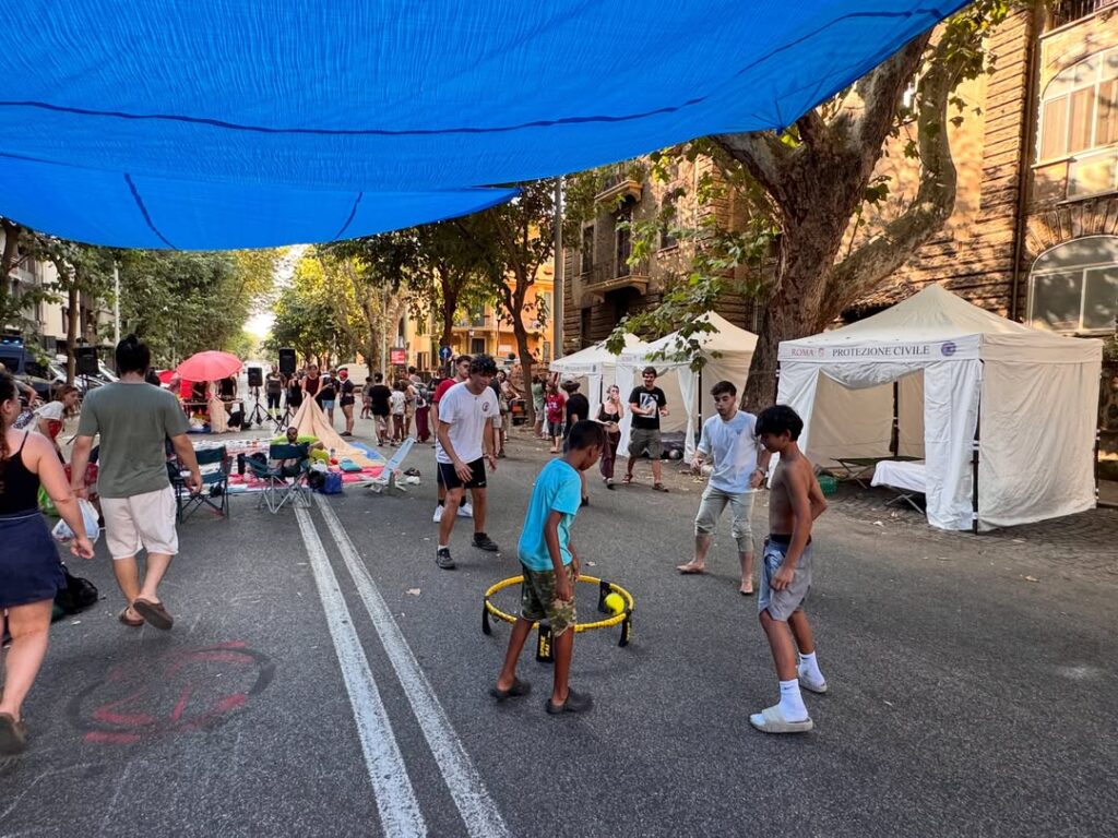 Street festival scene with a large blue canopy, tents, and people playing a ring-toss game on the road under the shade sail.